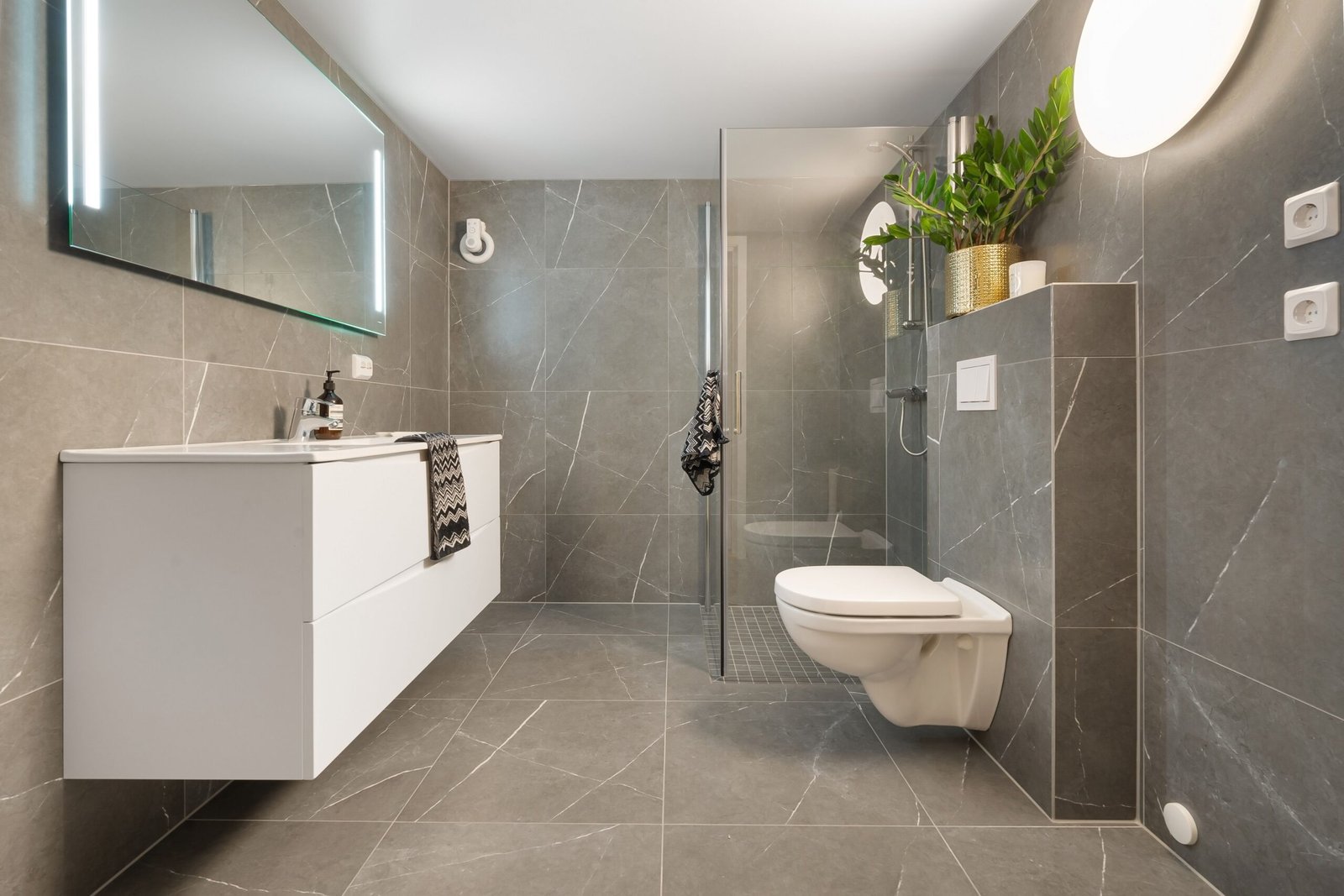 Stylish gray tiled bathroom featuring a shower, toilet, and illuminated mirror.