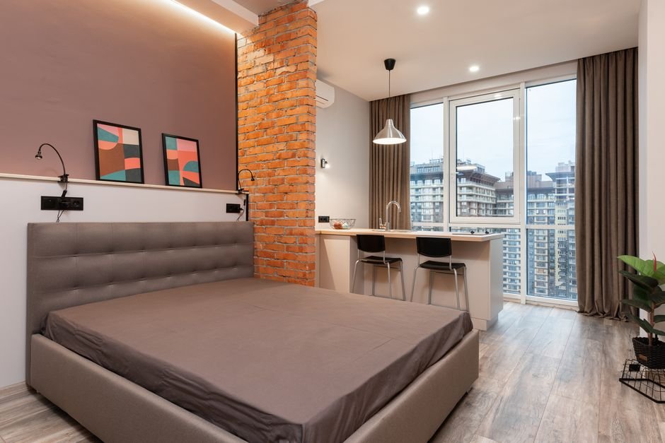 Modern light apartment interior with bed near counter with chairs near window with curtains and paintings on wall near potted plant
